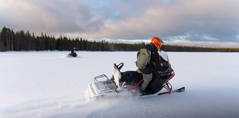 A snowmobile rider crouches low over the handlebars crossing open snow, another rider visible in the distance