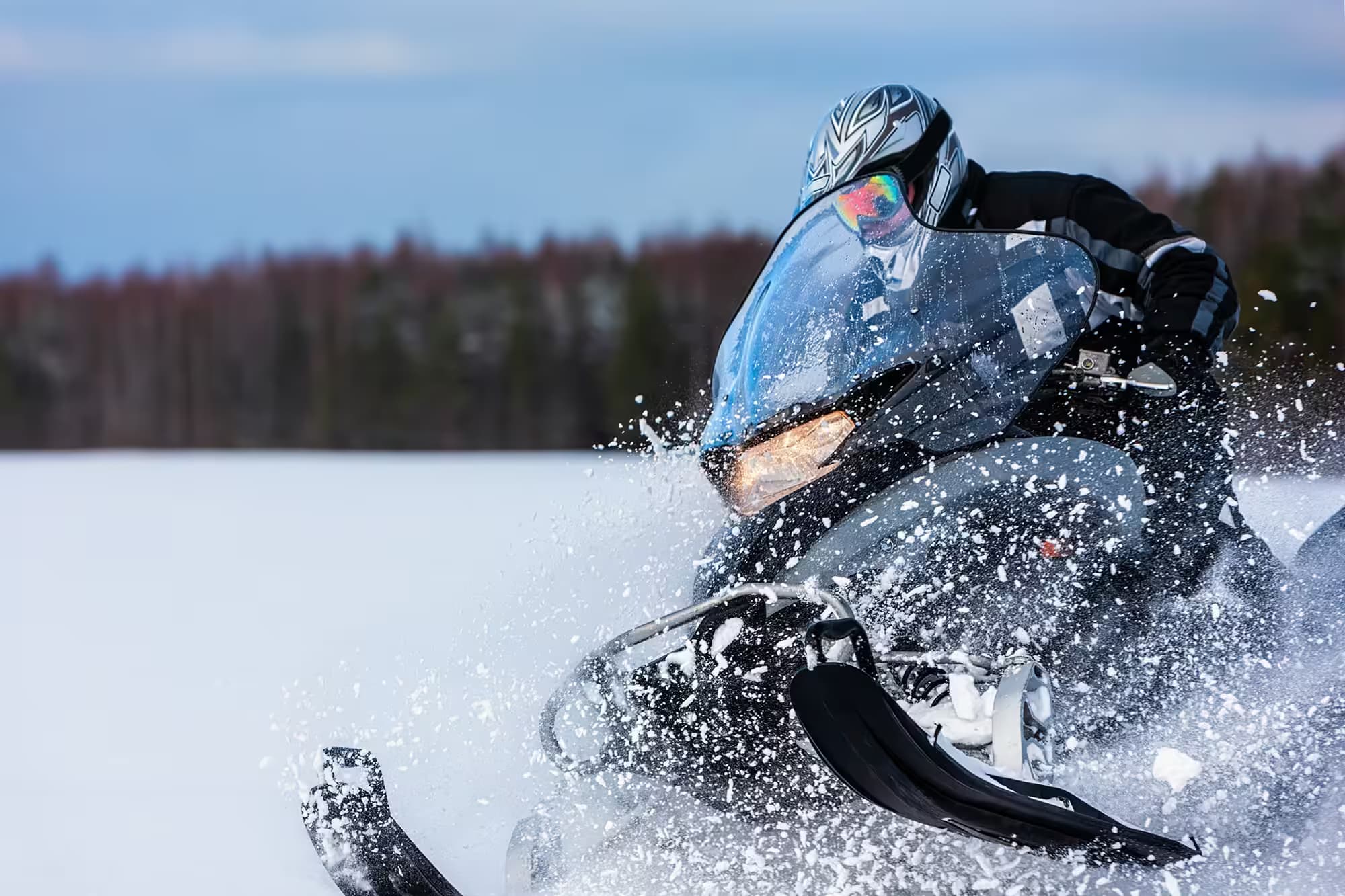 A helmeted snowmobile rider leans into a sharp turn, throwing up a wide spray of snow with a tree line behind