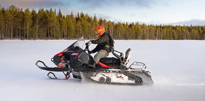 A snowmobile and its rider seen side-on, tracks curving away across an open field of snow