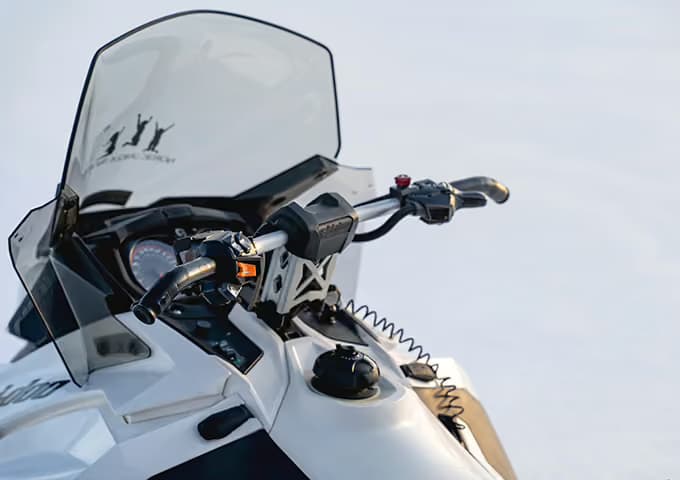 The cockpit of a white snowmobile — tall windscreen, handlebars and instrument dial — parked on open snow