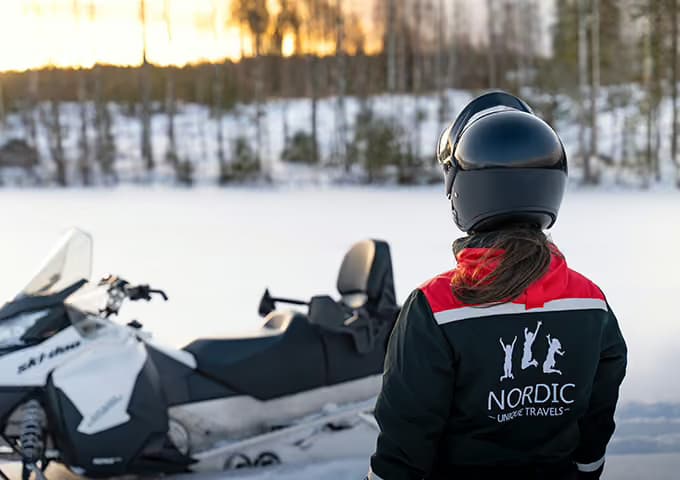 A rider in a helmet and dark guide's jacket looks out across a frozen lake at sunset, her parked snowmobile beside her