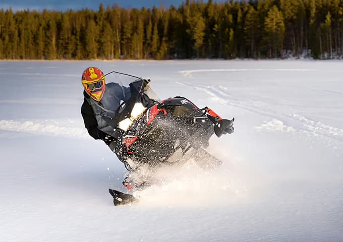 A rider leans hard into a turn on a red and black snowmobile, throwing up a wall of snow on a frozen lake