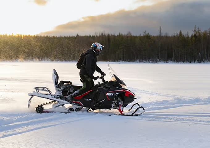 A rider stands on the footboards of a red and black snowmobile crossing a frozen lake at dusk