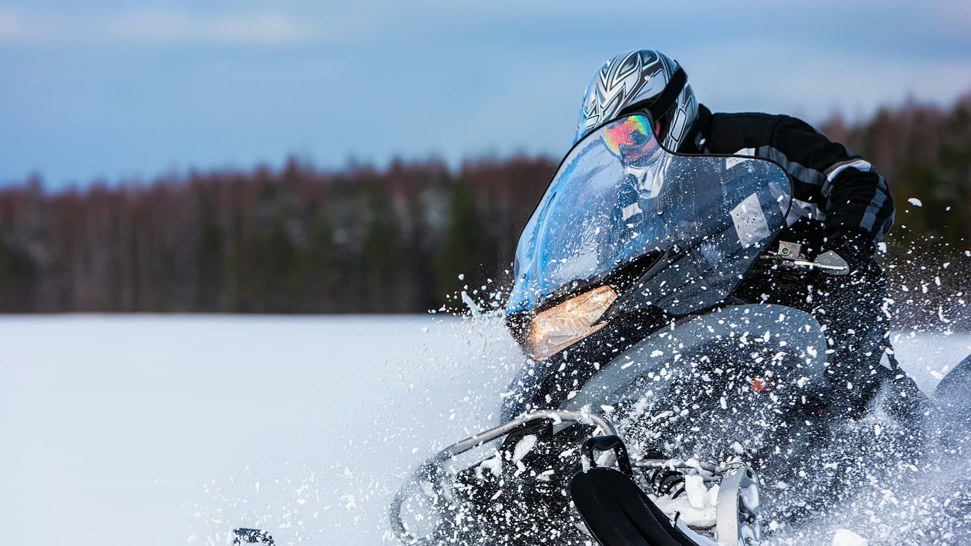 A helmeted snowmobile rider powers through deep snow, spray flying from the skis, a forest on the horizon behind