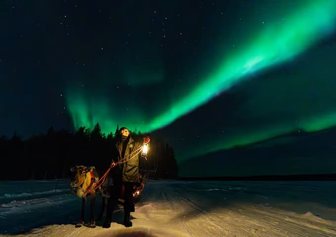A guide holding a lantern stands beside a reindeer on the snow beneath a bright green aurora