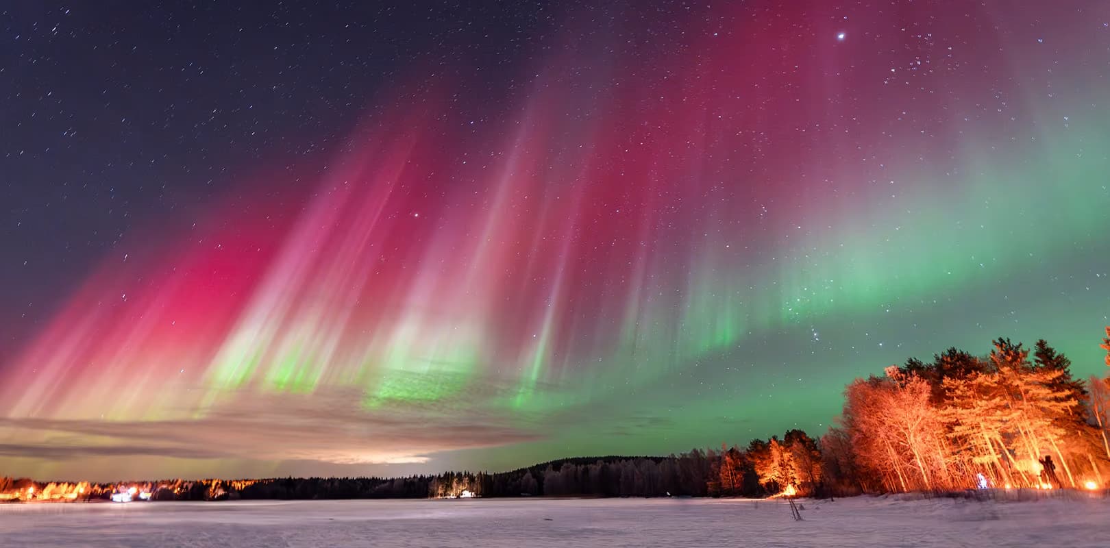 Wide bands of red and green aurora sweep across the sky above a snow-covered lake, lit trees and a bonfire on the shore