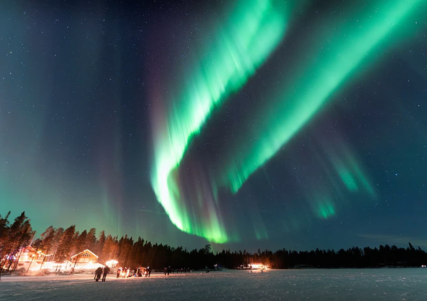 Green and violet aurora arcs stretch across the sky above a lit lakeside lodge and a group of guests on the ice at night